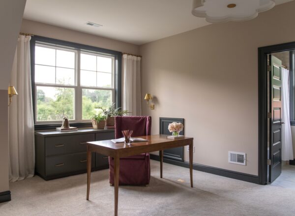 A home office with a wooden desk, maroon chair, potted plants, and large window with curtains. Light beige walls, dark trim, and an open door revealing another room with tiled flooring and greenery outside.