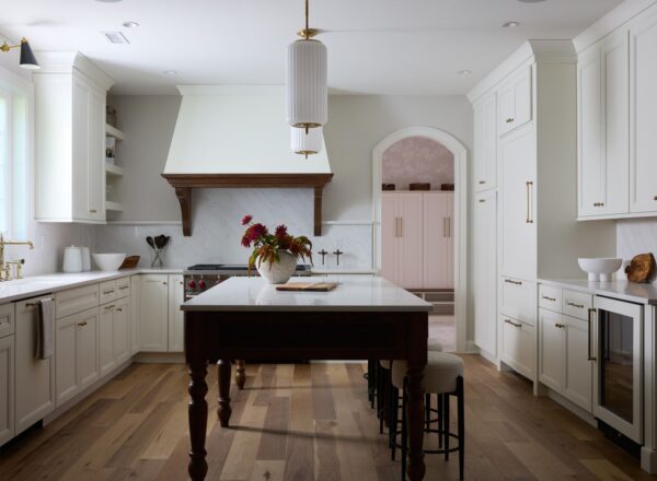Bright, modern kitchen with white cabinets, marble countertops, wood floor, and a large center island with stools. A vase of flowers sits on the island, and a window brings in natural light. An arched doorway leads to another room.