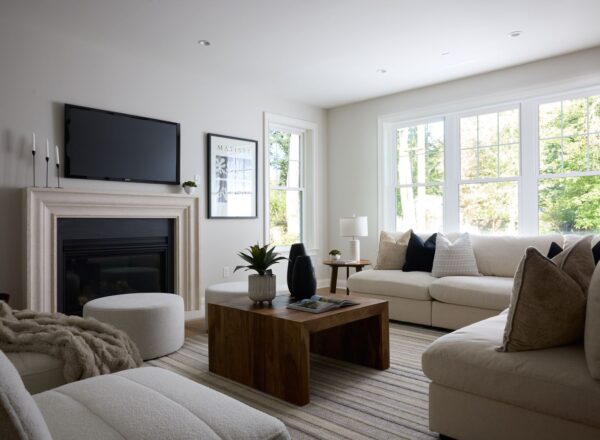 A bright, modern living room with a large window, white sofas, a striped rug, a wooden coffee table, and decorative accents, including candles and framed art above a fireplace with a television.