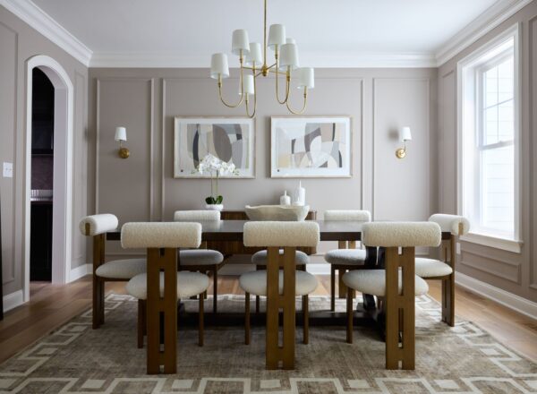 A modern dining room with a wooden table, eight cushioned chairs, abstract art on the walls, a gold chandelier, and large windows letting in natural light. Neutral tones and geometric decor create a sophisticated look.