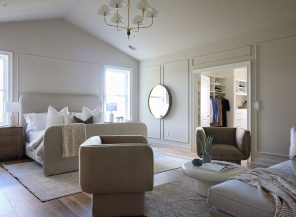 A modern, spacious bedroom with neutral tones, featuring a bed, armchairs, a round mirror, a chandelier, large windows, and an open walk-in closet with clothes and shelves. Light fills the room through the windows.