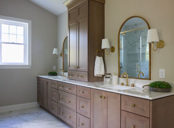 A spacious bathroom with wooden cabinets, double sinks, gold fixtures, two arched mirrors, white countertops, wall sconces, a towel ring, and a window letting in natural light. Marble floors and a glass shower are visible.