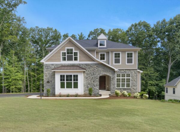 Two-story house with stone and siding exterior, large windows, gabled roof, and a neatly landscaped lawn, surrounded by trees under a clear blue sky.