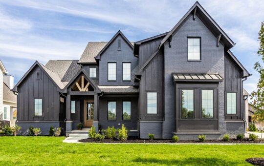 Modern two-story house with dark gray brick and siding exterior, large windows, peaked roofs, a small front porch with wooden accents, and well-kept green lawn and shrubs in front.