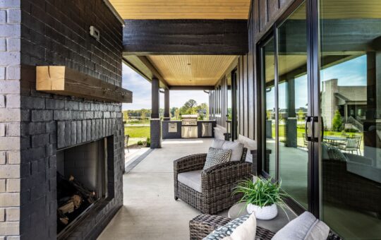 A covered patio features wicker chairs with cushions, a black brick fireplace, potted plant, and large glass doors. In the background, theres an outdoor kitchen and a view of a grassy yard.