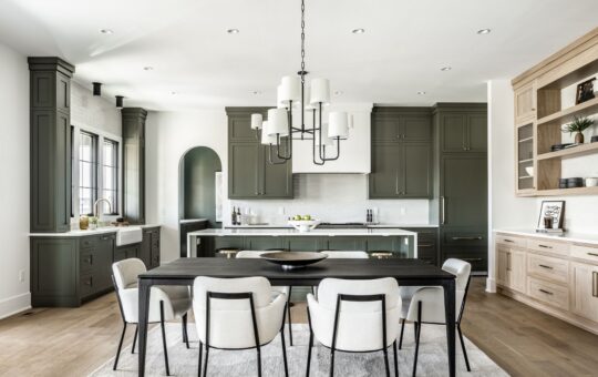 Modern kitchen and dining area with olive green cabinets, white countertops, light wood floors, and a black dining table surrounded by white chairs. Large windows and minimalist decor create a bright, airy atmosphere.