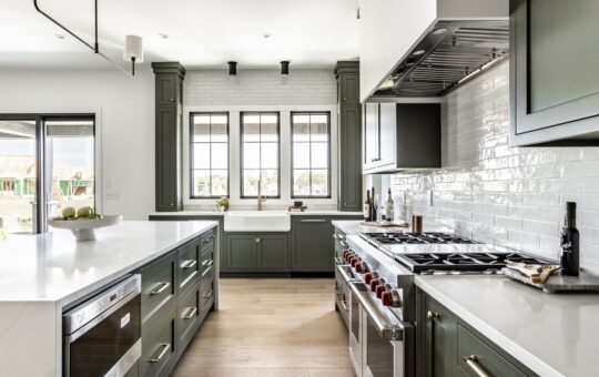 Modern kitchen with green cabinets, white countertops, stainless steel appliances, a farmhouse sink under three windows, light wood flooring, and white subway tile backsplash. Bowl of apples sits on the island.