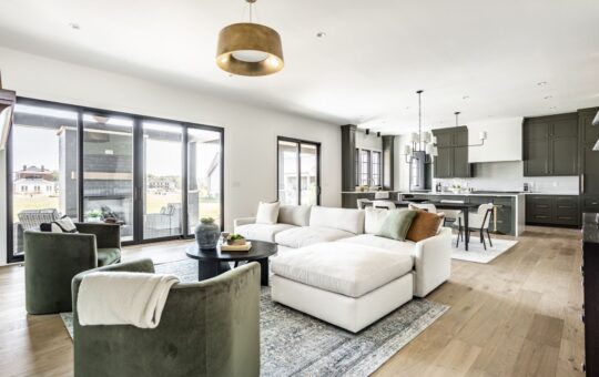 Modern open-concept living room and kitchen with large windows, green armchairs, white sectional sofa, area rug, dark cabinets, pendant lights, and natural wood flooring.