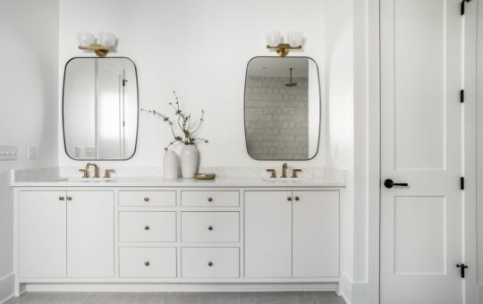 A modern bathroom with a double vanity featuring white cabinets, gold hardware, two rectangular mirrors, and two light fixtures. Two white vases with branches are placed between the sinks on a marble countertop.