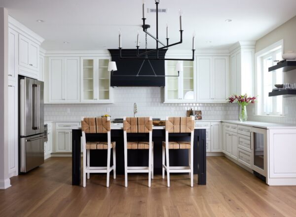 A modern kitchen with white cabinets, stainless steel appliances, a large island with four woven-back barstools, wood flooring, black light fixture, and a vase of pink flowers on the countertop near a window.