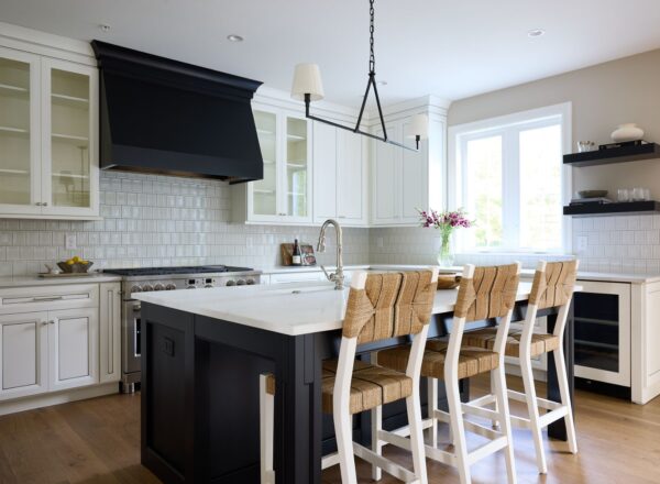 Modern kitchen with white cabinets, a black island, four wicker bar stools, a marble countertop, stainless steel appliances, tiled backsplash, large window, and vase of flowers on the island.