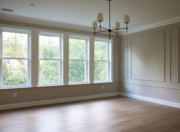 Bright, empty room with light wood flooring, beige walls, large windows letting in natural light, and a modern black chandelier hanging from the ceiling. Trim and molding line the walls, with a view of greenery outside.