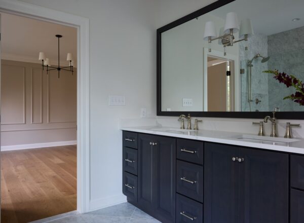 Modern bathroom with dark wood double vanity, large mirror, white countertop, dual sinks, brass fixtures, and wall sconces. An open doorway leads to a room with light wood flooring and a black chandelier.