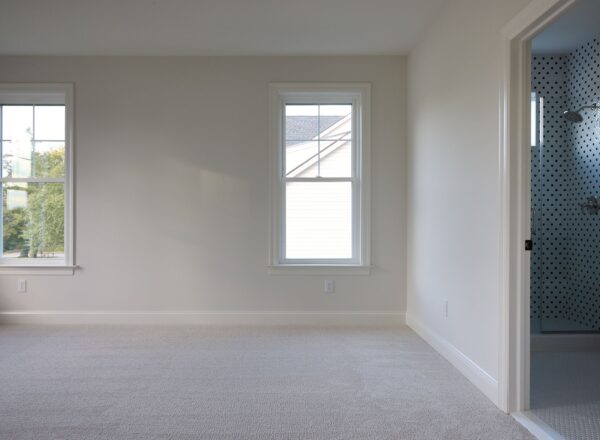A bright, empty room with two windows, carpeted floor, and white walls. An open door reveals a bathroom with a shower and white tiles with black polka dots.