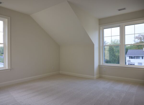 An empty, carpeted room with large windows letting in natural light, light-colored walls, a sloped ceiling, and views of trees and a neighboring house outside.