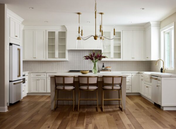 A modern kitchen with white cabinets, gold hardware, a large island with three chairs, wood flooring, pendant lights, a vase with flowers on the island, and sunlight coming through a window over the sink.