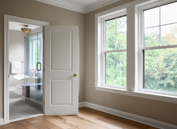 A bright, empty room with light wood floors and beige walls features two large windows and an open door leading to a modern bathroom with a glass shower, white tub, and gold fixtures. Green trees are visible outside.