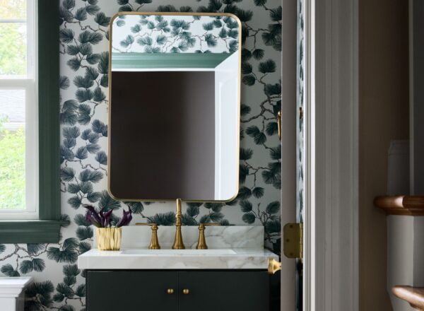 A stylish bathroom with pinecone-patterned wallpaper, a rectangular mirror with a gold frame, gold faucet, green vanity, marble countertop, and a window letting in natural light. A gold vase with dark flowers sits on the counter.