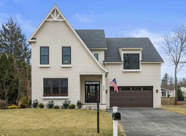 Two-story modern house with light brick exterior, dark-framed windows, and attached two-car garage. An American flag hangs near the front door. The house is surrounded by a lawn, trees, and a blacktop driveway.