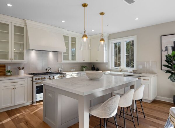 Modern kitchen with white cabinets, marble island with three cushioned stools, stainless steel appliances, pendant lights, wooden floors, large window, and decorative plants and artwork.