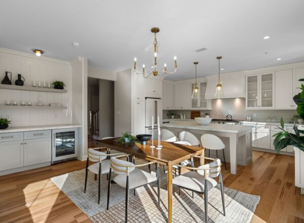 Bright, modern kitchen and dining area with white cabinets, an island, gold light fixtures, wooden dining table with six chairs, hardwood floors, and decorative plants. Natural light fills the space.