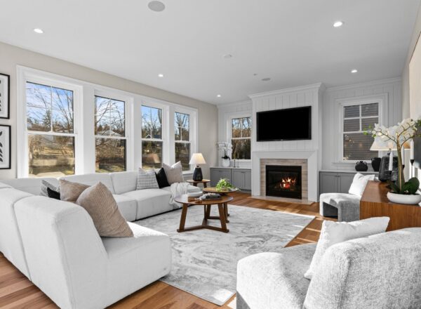 A modern living room with white sofas, a round coffee table, a fireplace below a mounted TV, large windows showing trees outside, and wooden floors with a light patterned rug.