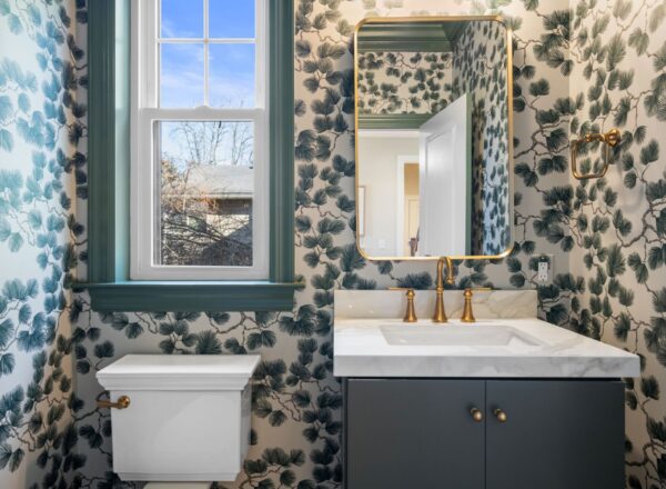 A bathroom with floral wallpaper, a window, a white toilet, a white countertop with a built-in sink, gold faucet and mirror, and black vanity. The window lets in natural light, and the decor has a modern, elegant style.