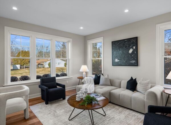 Modern living room with large windows, a light-colored sectional sofa, black chairs, a round wooden coffee table, white rug, and minimalist decor. Daylight streams in, showcasing a suburban neighborhood outside.