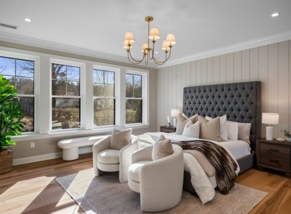 A modern, sunlit bedroom with large windows, a tufted gray headboard, neutral bedding, two round white chairs, wooden nightstands, a potted plant, a bench, and a gold chandelier.