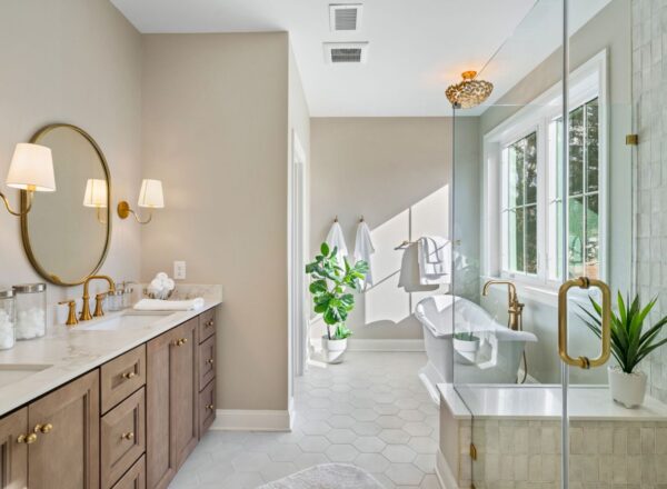 Modern bathroom with a double-sink vanity, round mirror, and gold fixtures. A glass shower is next to a soaker tub by large windows. White towels and plants add a fresh, elegant touch.