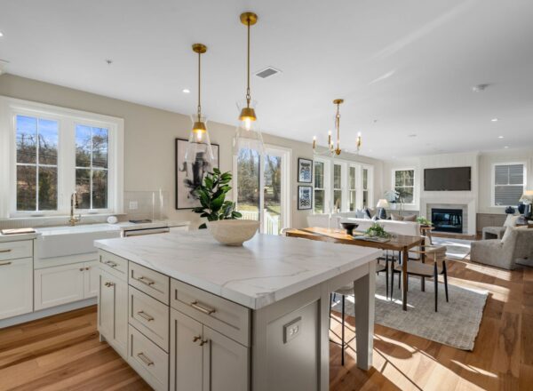 A bright, modern kitchen with white cabinets, marble island, gold pendant lights, and hardwood floors, opening to a dining area and cozy living room with a fireplace and large windows.