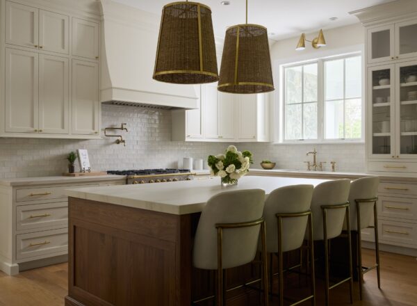 A modern kitchen with white cabinets, a large wood island with four upholstered stools, gold accents, two woven pendant lights, and a vase of flowers on the counter, with natural light from a window.