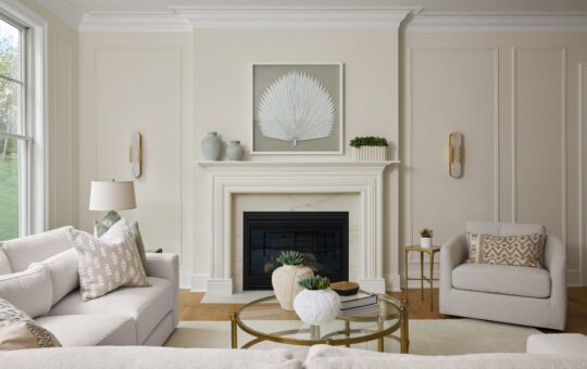 A modern living room with cream walls, a white fireplace, light-colored sofas, a glass coffee table with decor, and minimalist art above the mantel. Large windows bring in natural light.