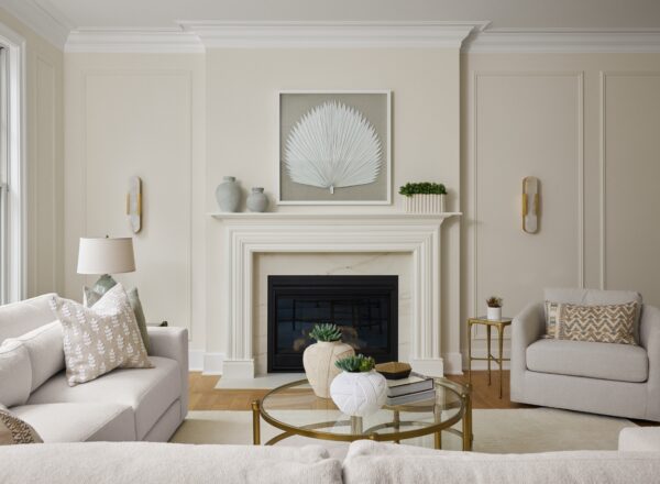 A modern living room with cream walls, a white fireplace, light-colored sofas, a glass coffee table with decor, and minimalist art above the mantel. Large windows bring in natural light.