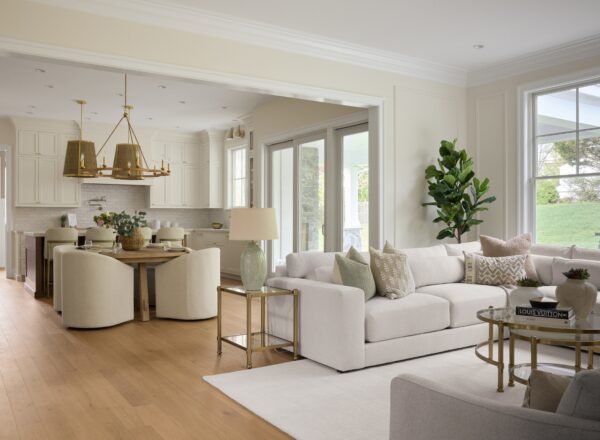 Bright, modern open-plan living room and kitchen with a large white sectional sofa, neutral decor, gold accents, a wooden dining table with six chairs, pendant lights, and large windows letting in natural light.