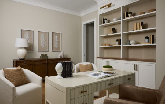A modern home office with a cream desk, two armchairs, built-in shelves with books and decor, framed botanical art, and neutral tones in the furnishings and walls.