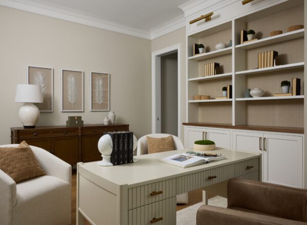 A modern home office with a cream desk, two armchairs, built-in shelves with books and decor, framed botanical art, and neutral tones in the furnishings and walls.