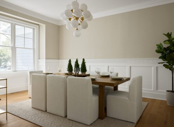 A modern dining room with a wooden table, six white upholstered chairs, a geometric rug, and a chandelier of round white bulbs. There are small potted trees on the table and a large window letting in natural light.