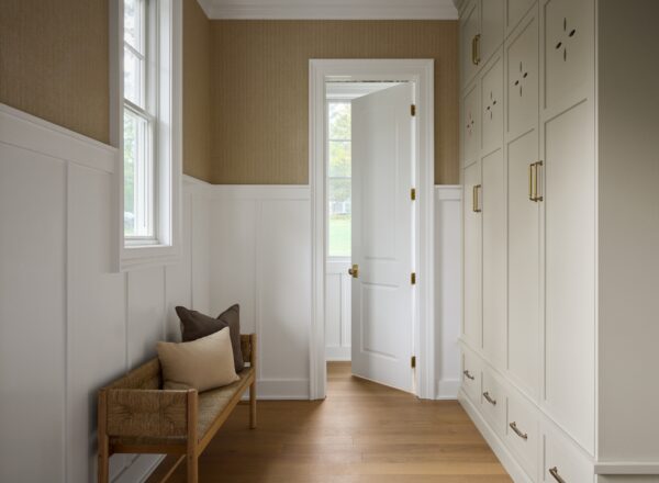 A bright hallway with a wooden bench topped with two cushions, built-in cream cabinets with brass handles, wooden floor, white wainscoting, and an open door revealing a green outdoor scene.