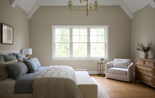 A cozy, modern bedroom with a neatly made bed, light bedding, a cushioned bench, an armchair by a window, a wooden dresser with a plant, and a gold chandelier hanging from a vaulted ceiling.