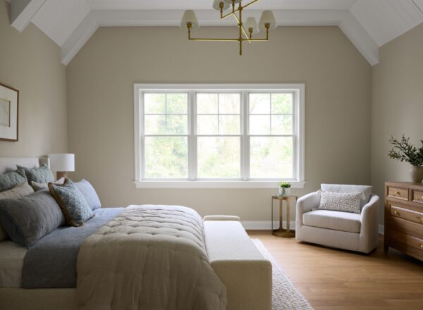 A cozy, modern bedroom with a neatly made bed, light bedding, a cushioned bench, an armchair by a window, a wooden dresser with a plant, and a gold chandelier hanging from a vaulted ceiling.