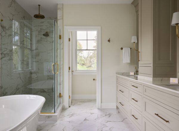 Modern bathroom with a freestanding tub, a glass-enclosed shower with marble walls, light cabinetry with gold hardware, marble floor tiles, a window, and neutral-colored walls.