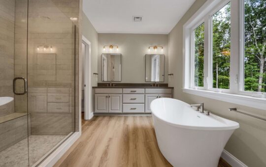 Modern bathroom with wood flooring, freestanding white bathtub by large windows, double vanity with two mirrors and sinks, and a glass-enclosed walk-in shower on the left. Natural light enters from the windows.