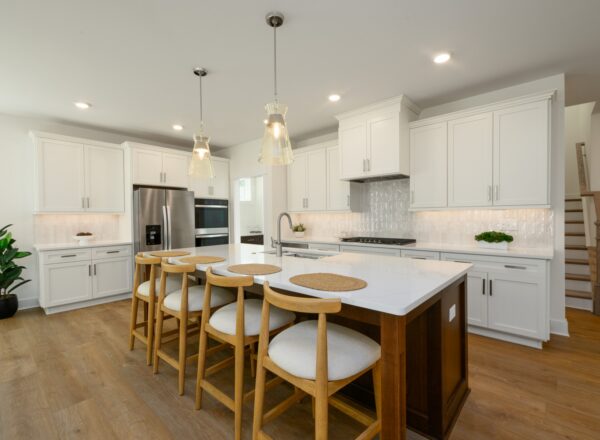 Modern kitchen with white cabinets, stainless steel appliances, a large island with a dark wood base, four cushioned bar stools, pendant lights, and light wood flooring. A staircase is visible in the background.