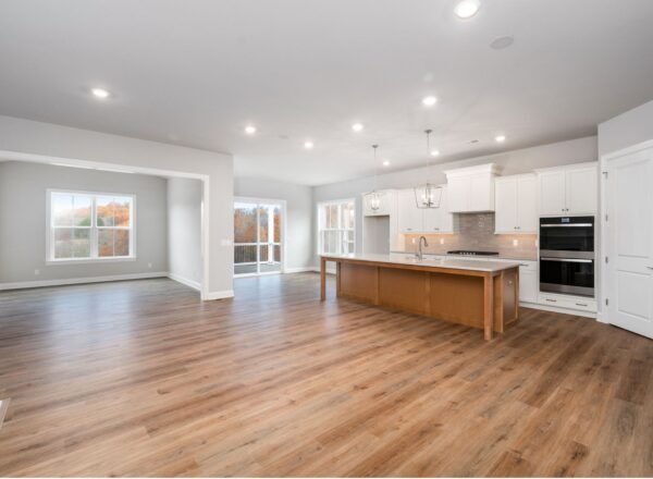 Spacious modern kitchen with white cabinets, a large island, stainless steel appliances, and wood flooring, open to a bright living area with large windows and neutral walls.