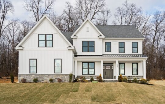 A two-story modern farmhouse with white siding, black trim, and a gray shingled roof, situated on a lawn with sparse landscaping and bare trees in the background.