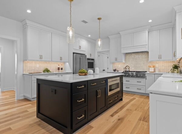 Modern kitchen with white cabinets, stainless steel appliances, a large black island with gold handles, light wood flooring, pendant lights, and decorative plants on the counters.