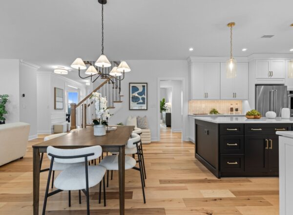 Modern open-concept kitchen and dining area with wooden floors, white cabinets, a black island, pendant lights, and a six-seat dining table. Stairs and decor elements visible in the background.