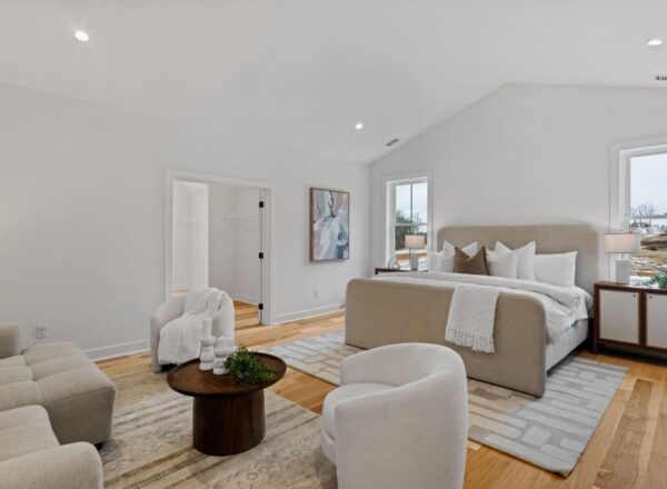 A spacious, modern bedroom with a beige upholstered bed, white bedding, two white armchairs, a light sofa, a round wooden coffee table, large windows, wooden floors, and minimalistic decor.