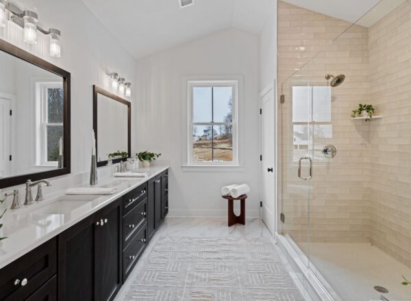 Modern bathroom with double sinks, dark cabinets, large mirrors, and wall lights. A glass-enclosed shower is on the right, with a small stool holding towels. A window lets in natural light, and the floor has a patterned rug.